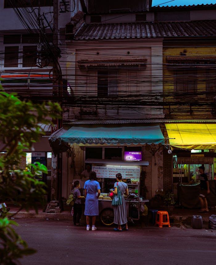 People gather at a food stall on a dimly lit urban street during twilight.