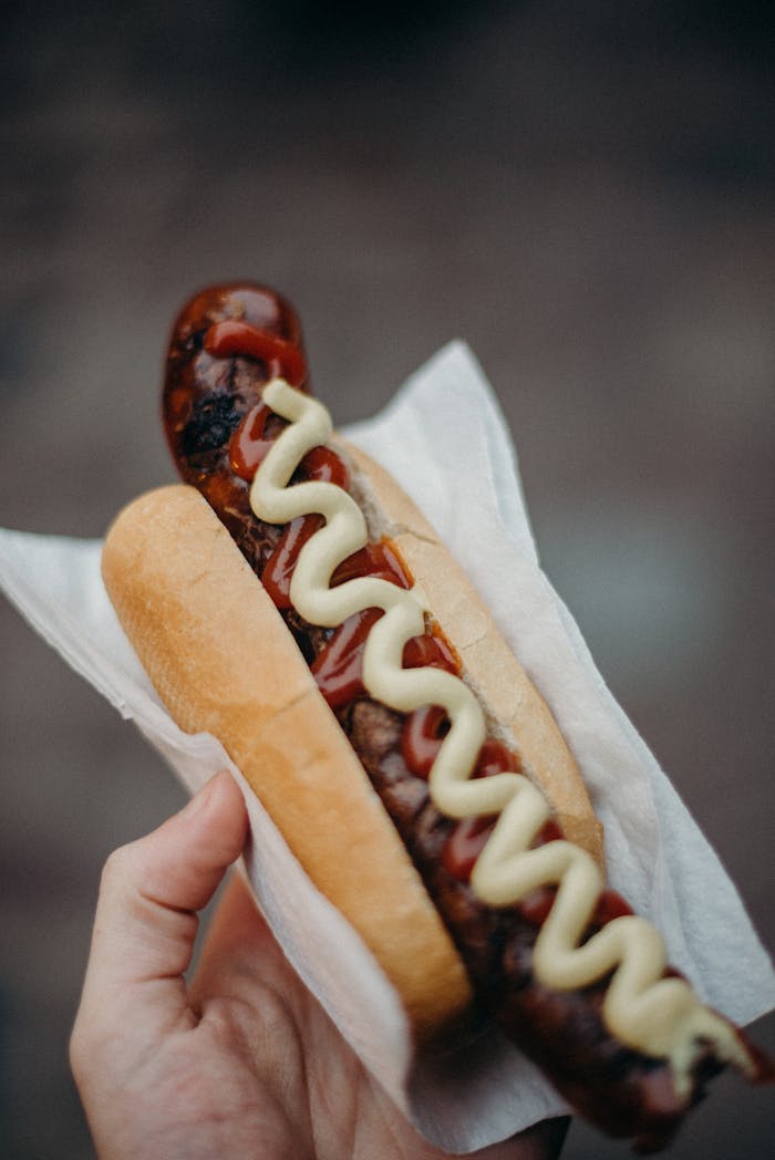 Close-up of a traditional German frankfurter hot dog held in hand, topped with mustard and ketchup.